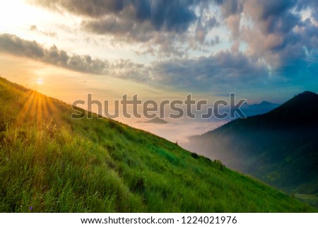 Mountain landscape in nice weather at sunrise. Green grassy steep hill, foggy valley and distant mountains under bright blue sky with lit by raising sun white clouds. Beauty of nature concept.