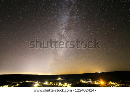 Beautiful starry dark night sky, Milky Way galaxy over rural landscape. Wide panorama, dark green hills, bright lights of road and distant buildings and glowing horizon. Copy space background.