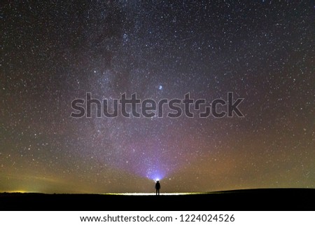 Back view of man with head flashlight standing on green grassy field under beautiful dark blue summer starry sky. Night photography, beauty of nature concept. Wide panorama, copy space background.