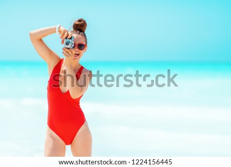 smiling modern woman in red beachwear taking photos with retro photo camera on the seashore