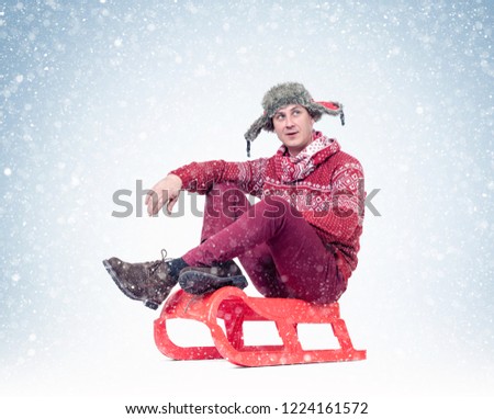 Happy man in a red sweater, scarf and hat is sitting on a sled looking at the falling snow.
