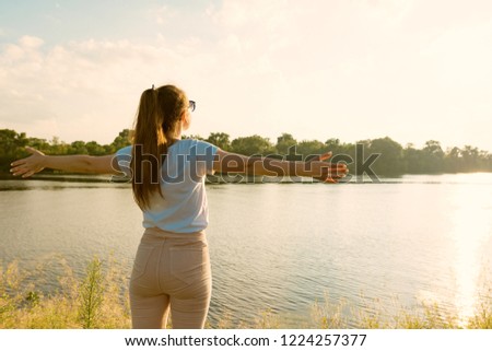 Young girl enjoys the sunset on the river beautiful landscape, the back of a girl spread her arms to the sides, golden hour.