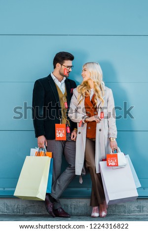 beautiful stylish couple holding shopping bags with sale tags and posing at blue wall