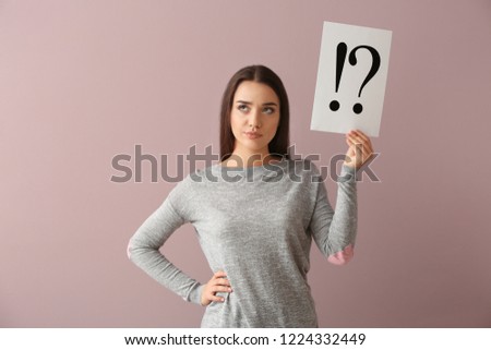 Thoughtful young woman holding sheet of paper with exclamation and interrogation marks on color background