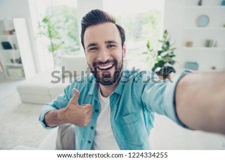 Careless carefree cheerful positive brunet hair man stand in light studio apartment in casual blue shirt take picture on smart phone cellular make beaming white smile give thumb up