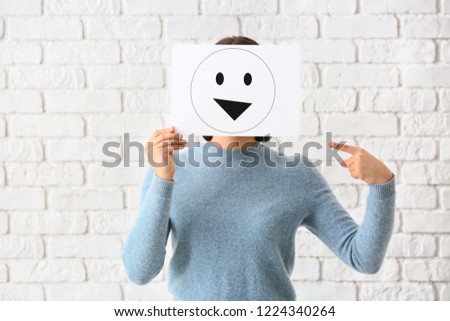 Young woman hiding face behind sheet of paper with drawn emoticon against white brick wall