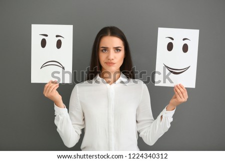 Emotional woman holding sheets of paper with drawn emoticons on grey background