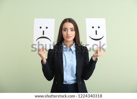 Young woman holding sheets of paper with drawn emoticons on light background