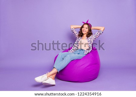 Full size portrait of attractive nice lady with her hairdo she look camera make white beaming smile hold hands behind head sit isolated on purple background in checkered shirt denim casual jeans
