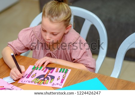 Needlework, crafts children.Little girl create a greeting card image of the Jewish holiday of Hanukkah. Kid pastes stickers - candles and drawing menorah, dreidel, oil jar on paper on wooden table.