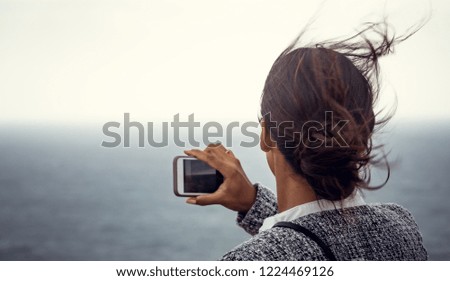 Beautiful young woman taking photos and selfies with her smartphone of the ocean with wind going through her long hair.