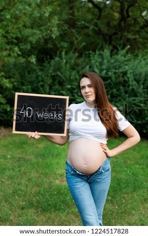 Beautiful smiling, stylish pregnant woman in jeans and checked shirt holding black board in hands. Brunette mom emotionally posing in park. Mother with belly spending time outdoors. Weeks before birth
