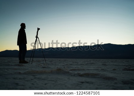 Male silhouette of photographer standing near tripod having expedition in death valley in twilight, man using professional equipment for making night shooting of wild nature and landscape in desert