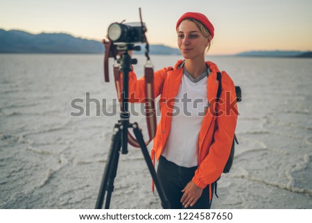 Positive skilled woman making settings on camera waiting for sunset for shooting video in desert during journey, professional female photographer taking pictures of nature and landscape via tripod