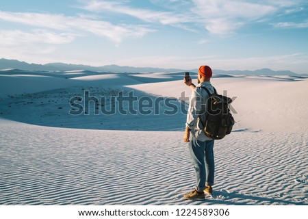 Hipster guy wanderlust making selfie on smartphone camera on breathtaking scenery of desert back view of male traveler with rucksack using telephone for making picture during journey to White sands 
