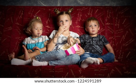 Children's movies: Three children watch movies at home on a big red sofa in the dark and eat popcorn.