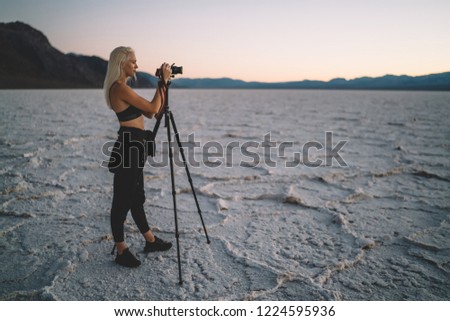 Young blonde caucasian woman shooting video on camera with tripod during evening twilights in death valley, hipster girl wanderlust making photos on professional equipment  on expedition trip