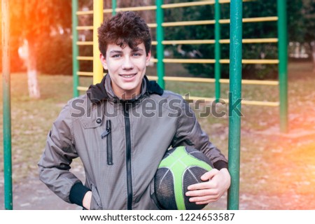 Happy teenager in a jacket and a basketball ball in his hand smiles at the background of a sports ground near the school. The teen has braces for teeth equalizing