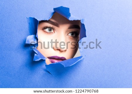 The face of a young beautiful girl with a bright make-up and puffy purple lips peers into a hole in violet paper.
