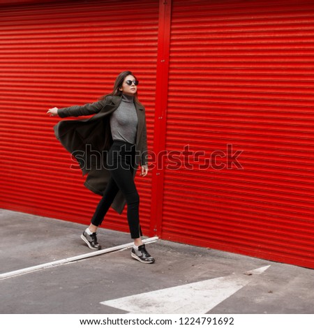 Stylish beautiful young woman with sunglasses in a fashion green coat walks on the street near the red metal gates.