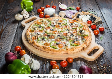 Big pizza with cheese, tomatoes, black olives and paprika on a round cutting board on a dark wooden background. Ingredients. Royalty-Free Stock Photo #1224804598