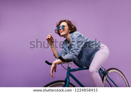 Carefree short-haired lady sitting on bicycle in studio. Happy caucasian girl with wavy hairstyle expressing positive emotions.