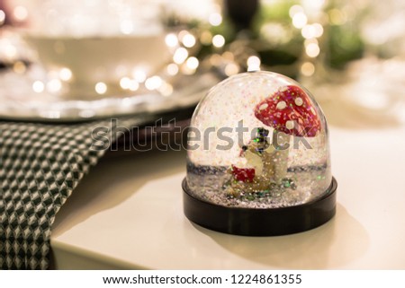 Magic Snow Ball with a little fox reading a book sitting under a mushroom. Snow Globe on Christmas table. Holiday Party Decorations, moody shallow depth of field image with bokeh Christmas lights.