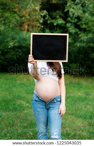 Beautiful stylish pregnant woman in jeans and checked shirt holding black board in hands. Brunette mom emotionally posing, hiding face. Mother with belly outdoors in park. Weeks before birth of baby.