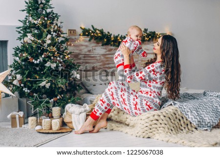 Happy mom with her little daughter in holiday clothing with printed deers and snowflakes having fun on the bed in cozy room with a Christmas tree and Christmas lights. New year and Christmas concept.
