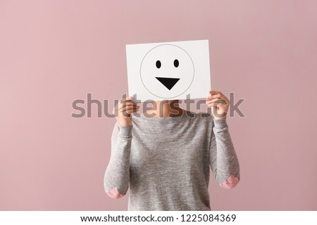 Young woman hiding face behind sheet of paper with drawn emoticon on color background