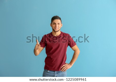 Young man in stylish t-shirt showing thumb-up gesture on color background