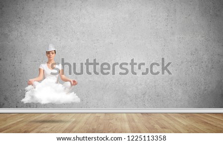 Young woman keeping eyes closed and looking concentrated while meditating on cloud in the air with gray concrete wall on background.