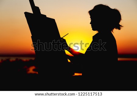 silhouette of a young woman painting a picture with paint brush and palette on canvas on an easel, girl face profile engaged in art on the nature in a field at sunset