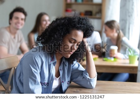 Sad mixed race girl sitting alone separately from other mates feels unhappy. Friends having fun together in cafe ignoring or scoffing at african peer. Outcast, discrimination and teen problem concept Royalty-Free Stock Photo #1225131247