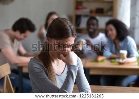 Diverse multi-ethnic friends sitting together in cafe talking having fun, focus on frustrated shy girl sitting separately by others teenagers feels unhappy because peers not accept her she is outcast Royalty-Free Stock Photo #1225131265