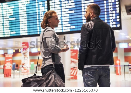 Discussion about planning a trip. Photo of two comrades situating in airport near flight information display system.