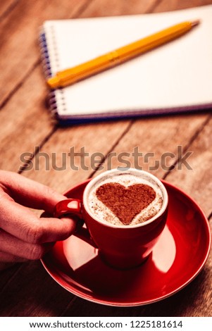 female hand holding cup of coffee with cream heart shape symbol near notebook and pen on wooden table. Above view