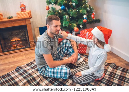 Lovely picture of girl touching nose of her father with finger. She looks at him and smiles. Young man in pajama looks at daughter. He smiles too. They sit in decorated room.