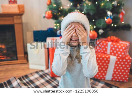 Lovely picture of small girl sitting on knees and covering eyes with hands. She smiles. Girl is positive. She wears white costume and hat. There are Christmas tree with toys under it behind her.