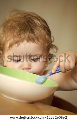 Portrait of serious little cute male kid with blonde curly hair and round cheecks eating from green plate with spoon closeup, vertical picture