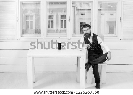 Hipster sitting on wooden chair at table with bottle and glass of red wine. Brutal man with beard and moustache indoors on white window background. Alcohol and convive. Unhealthy lifestyle. Bad habits