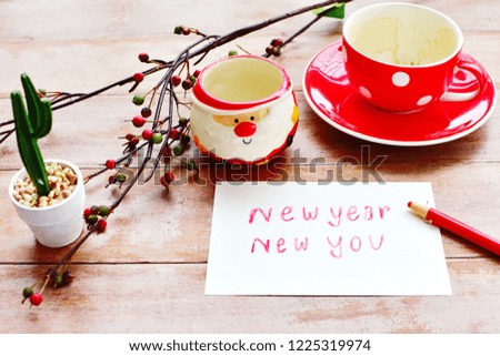 Drink coffee finished and write letter words New Year new you in white paper, red cup without drink, Santa claus cartoon cup, cactus in white pot, branch of berry on old wood board, Holidays concept 
