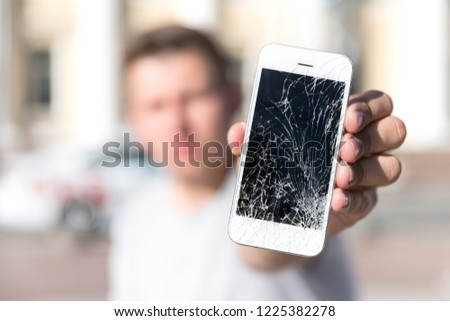 Caucasian young male holding and showing broken smartphone screen (display), selective focus on smartphone, blurred face, outdoors. 