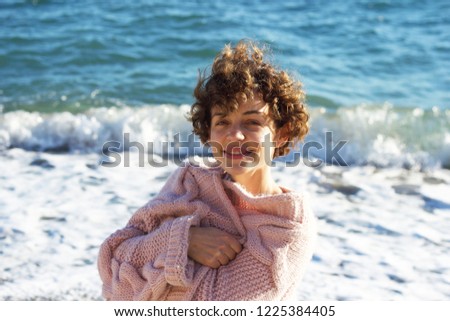 An adorable, smiling, curly-haired,  short-haired brunette in a rose sweater on the beach