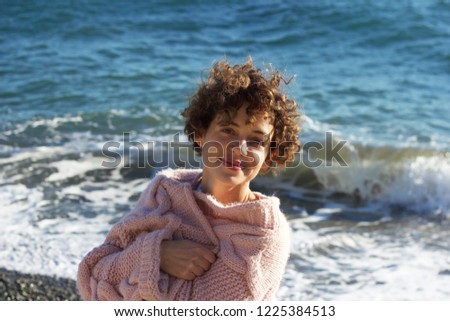 A dazzling, smiling, curly-haired,  short-haired brunette in a rose sweater on the beach Royalty-Free Stock Photo #1225384513