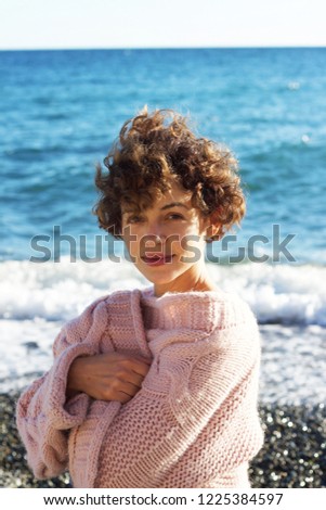 A sunny, smiling, curly-haired,  short-haired brunette in a rose sweater on the beach Royalty-Free Stock Photo #1225384597