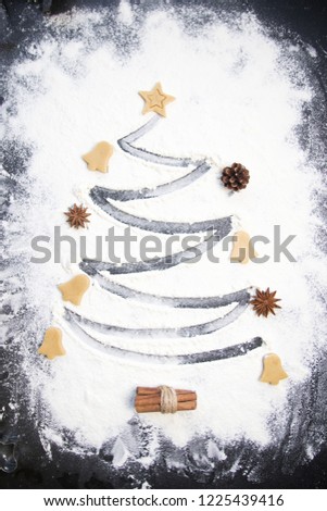 Christmas cooking: fir tree made from flour on a dark table, ingredients for baking and dried fruits on dark background, top view