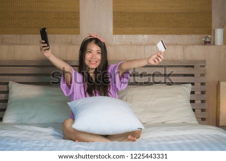 young beautiful and happy Asian Japanese woman at home bedroom using credit card internet banking with mobile phone in bed smiling cheerful in e-commerce business and online shopping