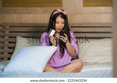 young beautiful and happy Asian Korean woman at home bedroom using credit card internet banking with mobile phone in bed smiling cheerful in e-commerce business and online shopping