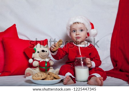 A little kid in a red cap eats a cookies and milk. Christmas photography of a baby in a red cap. New Year holidays and Christmas.
